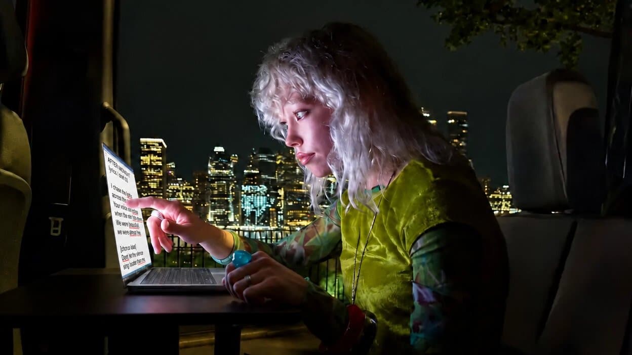At night in a parked van, a writer touches a laptop screen to select a line of text with typos underlined in red.